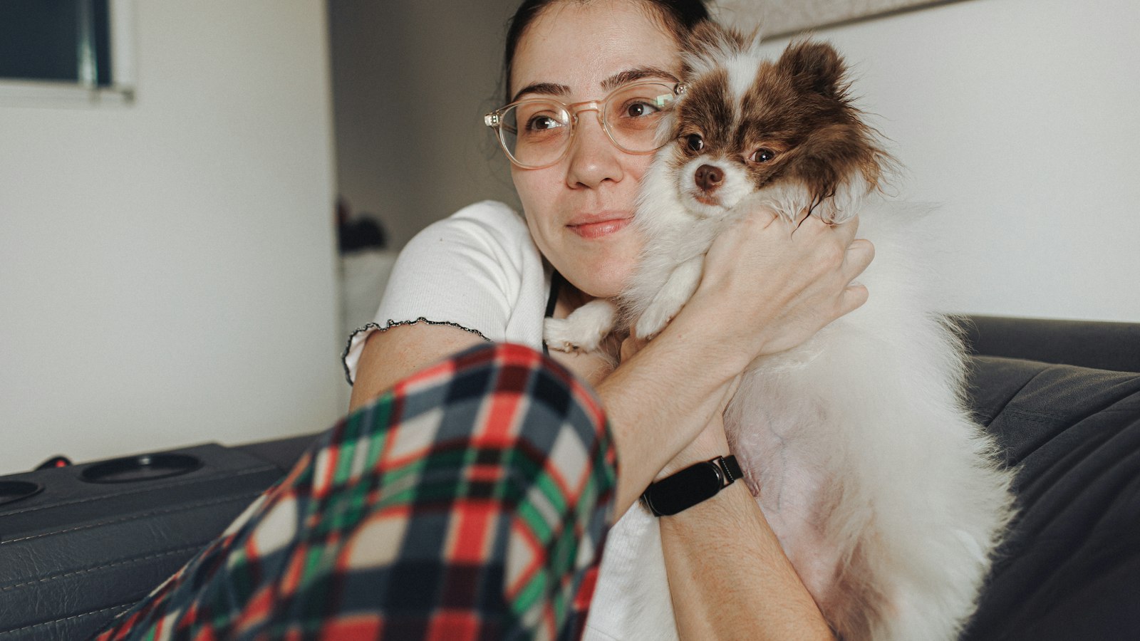 A woman in glasses sits on a couch holding a small white and brown dog close to her face